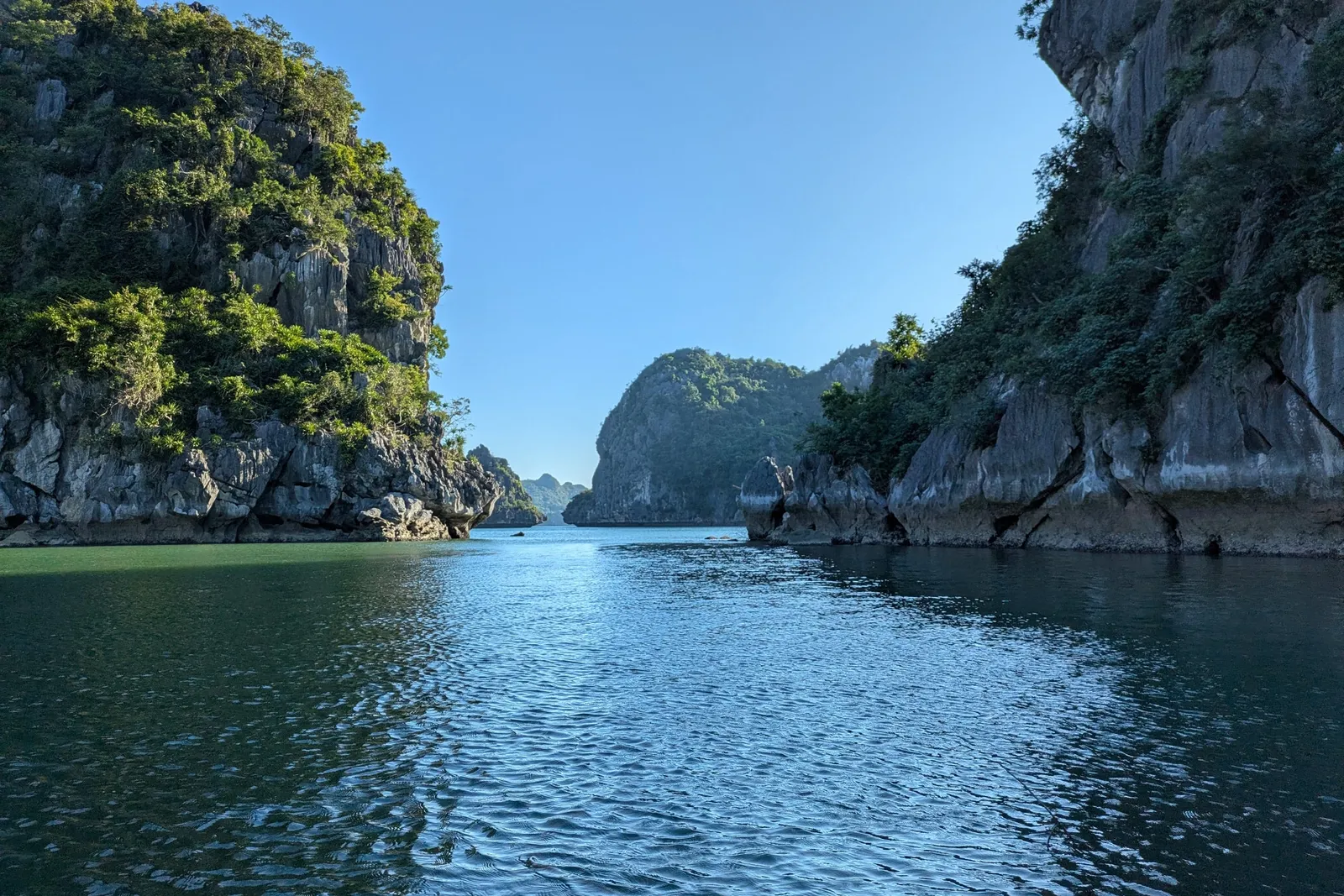 Calm water channel between tall limestone karst formations covered in vegetation under blue sky.