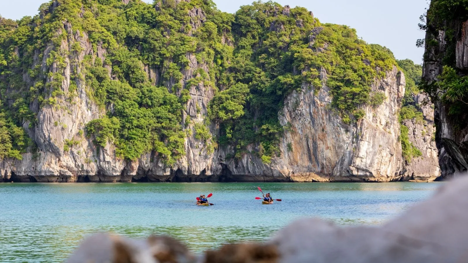 Two tandem kayaks with paddlers on calm turquoise water in front of tall green-covered limestone karst cliffs, with blurred rocks in the foreground.