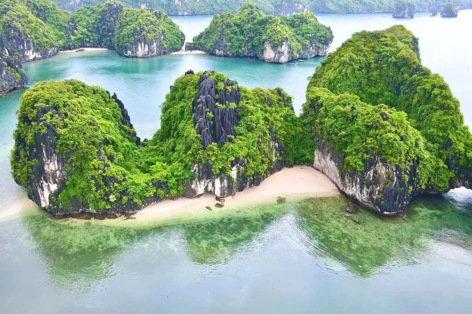 Aerial view of green-covered limestone karst islands with a small white sand beach and turquoise-green shallow water.
