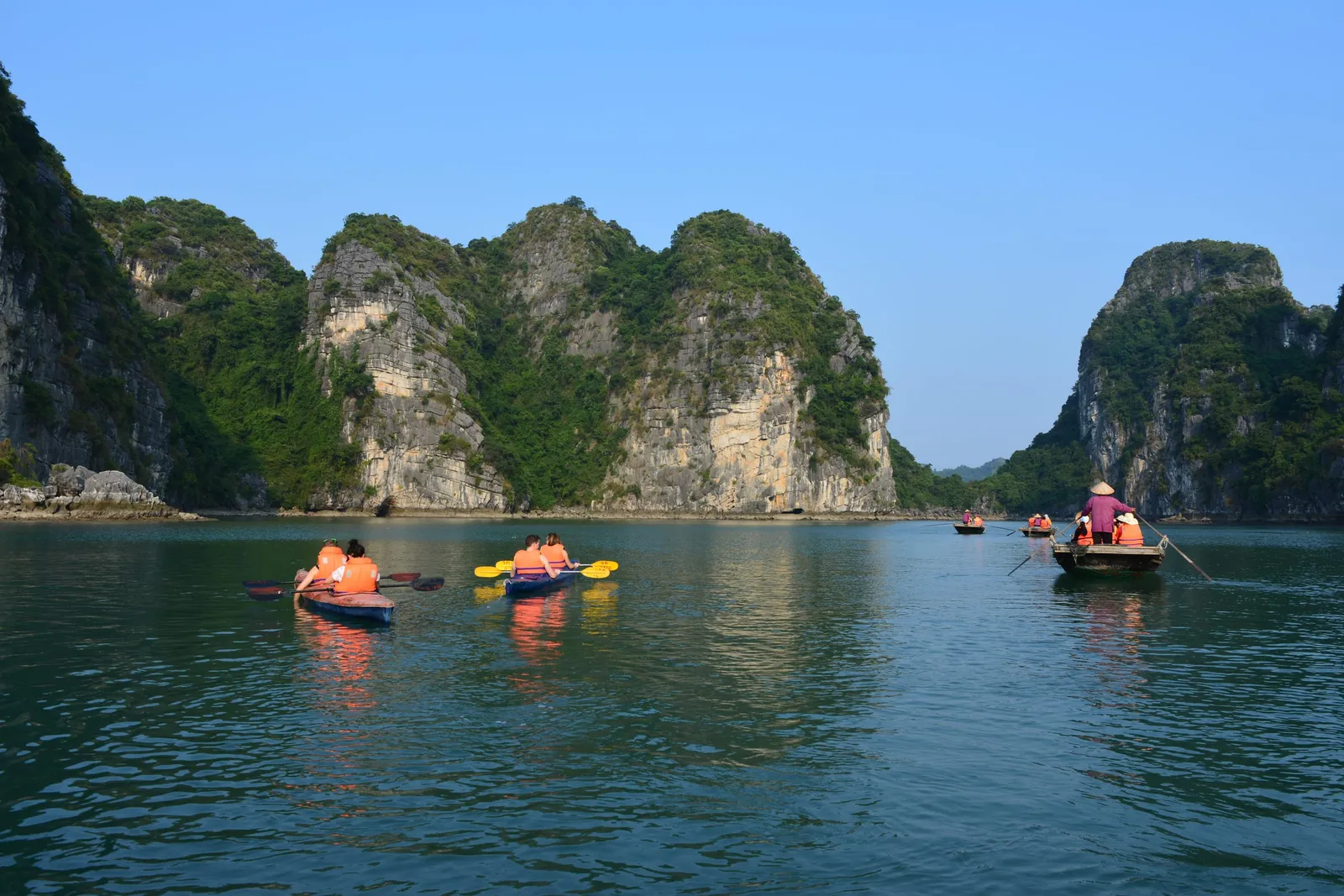 Kayakers in orange life jackets and a rower in a conical hat in small boats on calm green water in front of forested limestone karst cliffs under a clear blue sky.