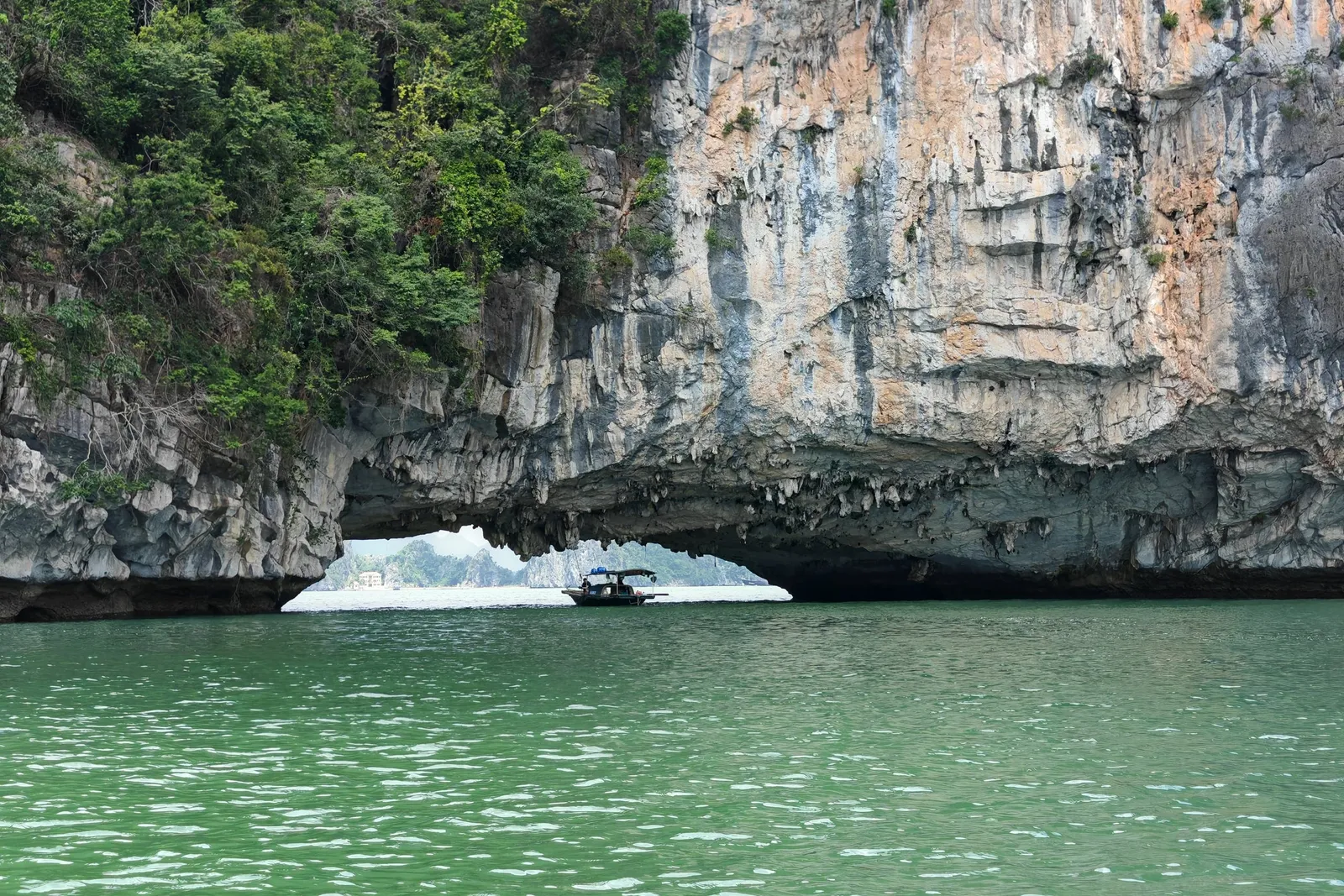 A limestone karst with a natural arch over green water with a small boat passing through the opening.