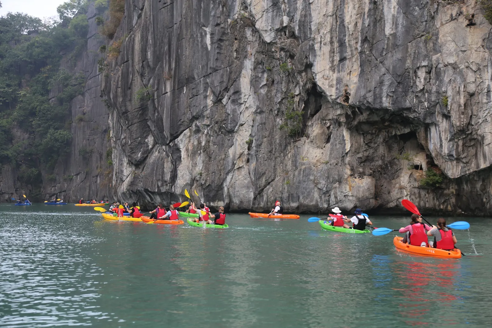 Multiple kayakers in colorful kayaks paddle near towering limestone cliffs with a cave opening visible in the rock face.