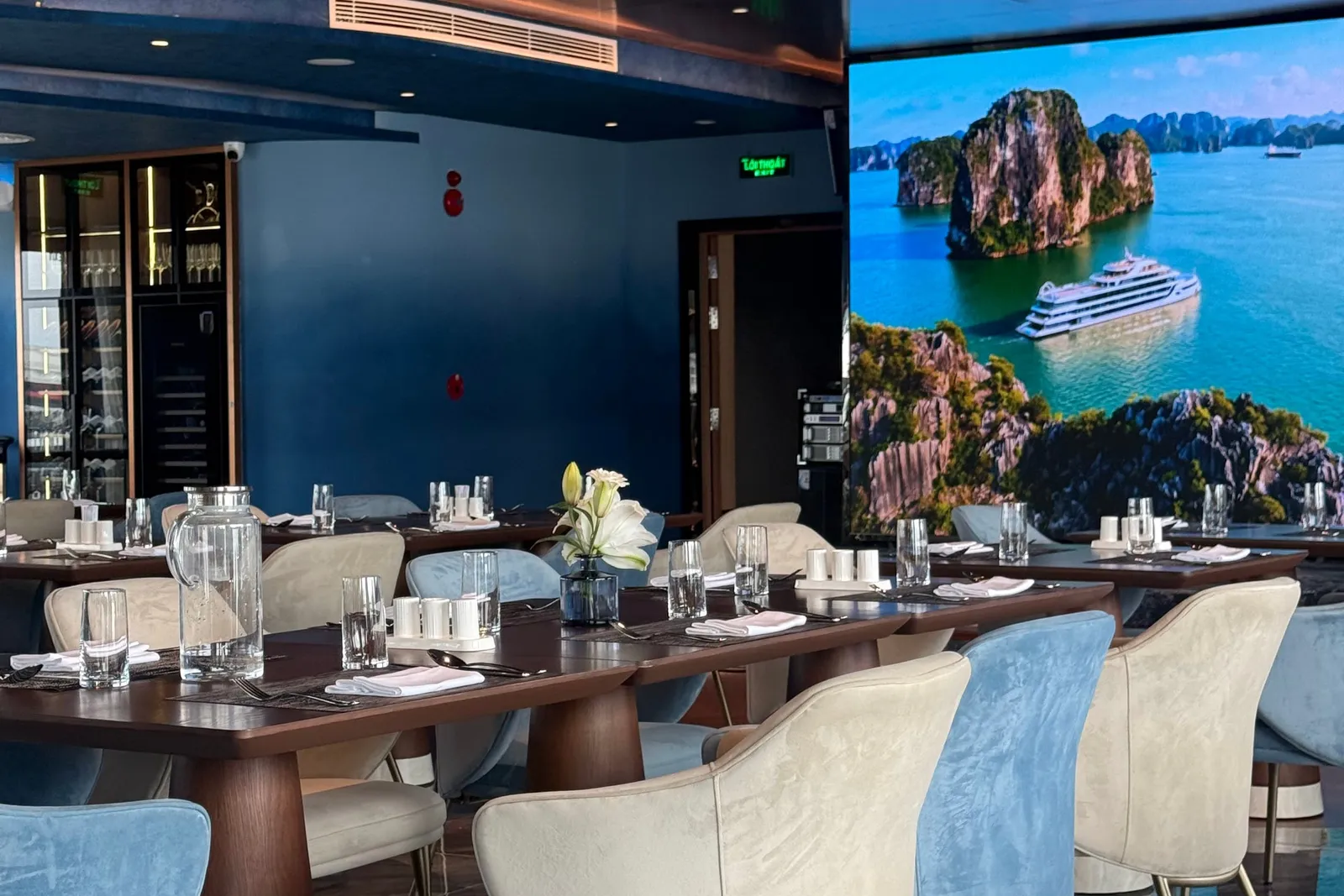 An indoor dining room with set wooden tables and blue and cream velvet chairs, with a large wall screen showing a cruise ship among limestone karsts in a bay.