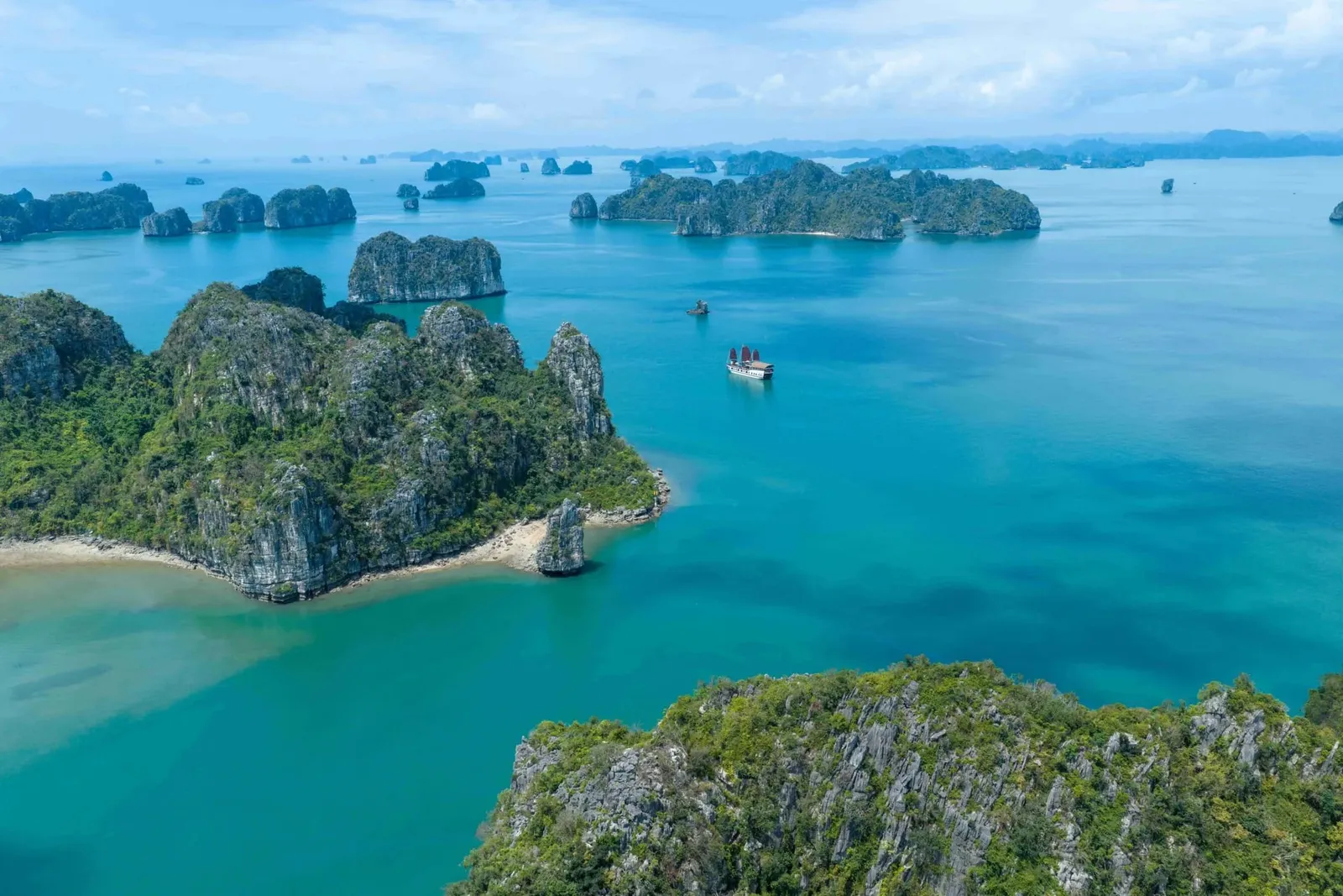 An aerial view of turquoise water dotted with green limestone karst islands, with a single small junk boat with red sails in the middle distance.