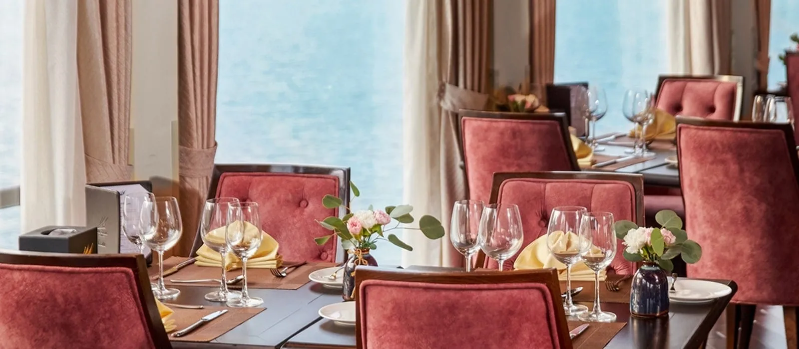 A dining room with set tables, pink velvet chairs, wine glasses, yellow napkins and small flower vases, with large windows showing calm blue-grey water outside.