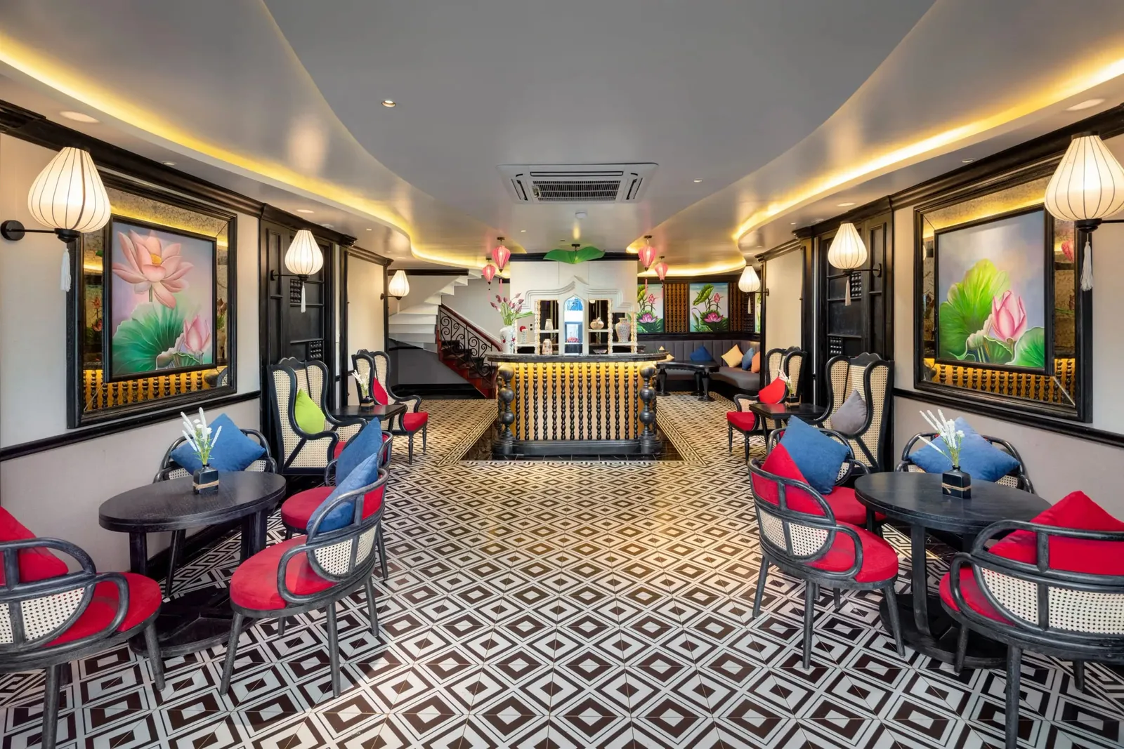 A lounge interior with black-and-white geometric tile flooring, black rattan chairs with red cushions, round dark tables, lotus paintings, lantern wall lamps, a central bar and a staircase at the back.