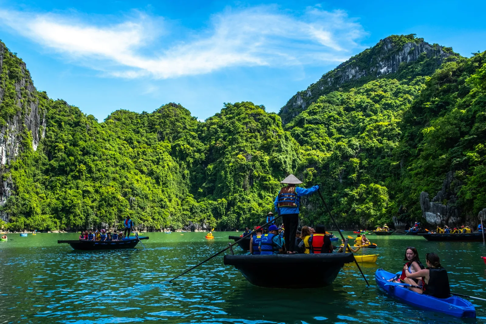A rower in a conical hat stands on a small black rowing boat carrying life-jacketed passengers, with kayaks and other boats on green water in front of steep limestone karst hills covered in dense greenery under a blue sky.