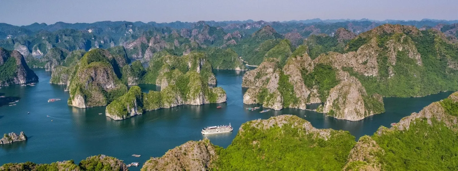 Aerial panorama of green limestone karst islands in a blue bay with a white multi-deck cruise ship in the centre and a few small boats scattered around.