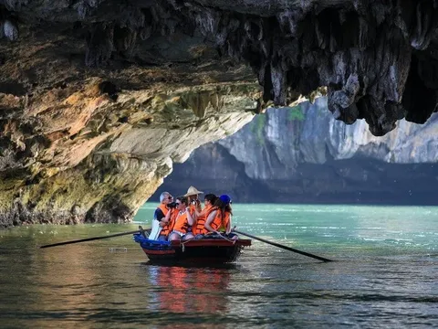 A group of tourists in orange life jackets sit in a small rowboat inside a limestone cave with turquoise water and dramatic rock formations.