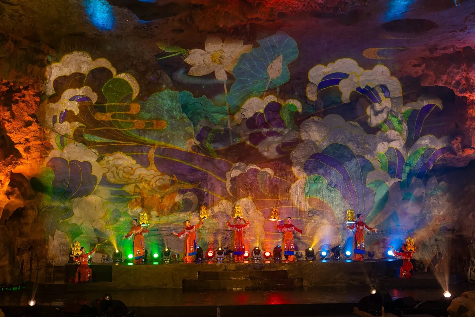Performers in orange-red costumes holding small golden pagoda lanterns on a stage inside a cave, with a lotus-pattern light projection on the rock wall and colored stage spotlights in front.