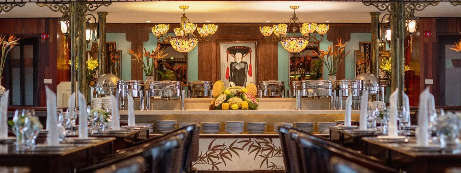Interior of a restaurant dining room with set tables, a buffet counter with chafing dishes and a fruit display, Tiffany-style pendant lamps, and a framed portrait of a woman in traditional Vietnamese dress.