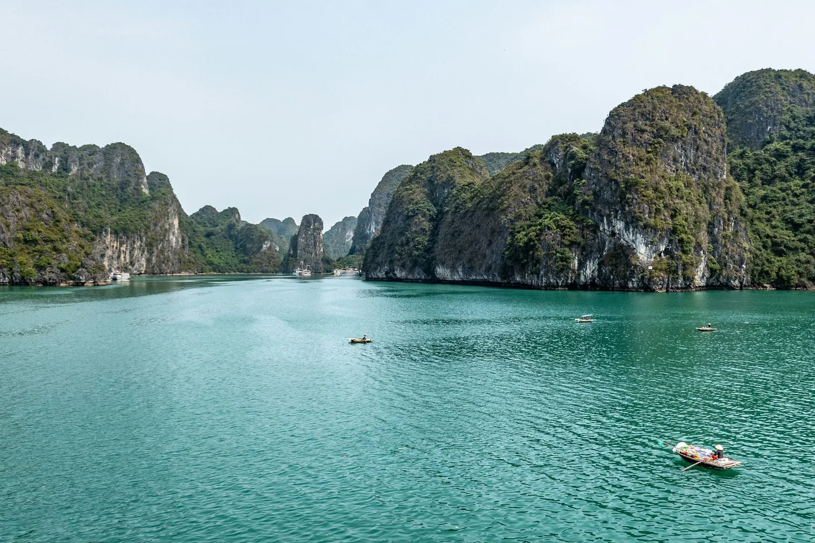 Turquoise bay water framed by tree-covered limestone karst islands, with a few small rowing boats, one carrying colorful goods, and distant larger boats under a hazy sky.