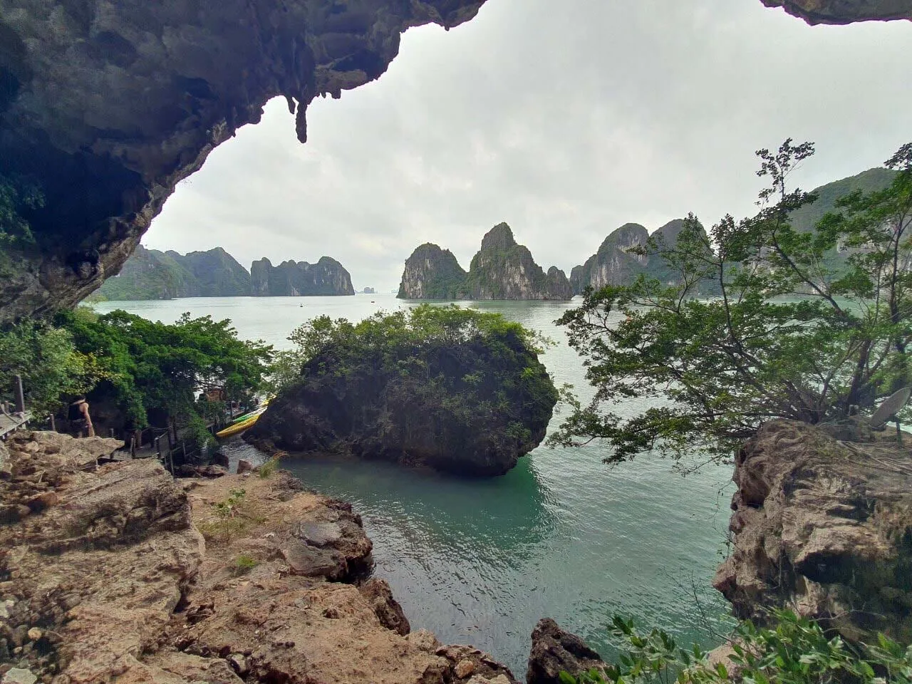 A view from inside a cave looking out at limestone karst islands rising from green water with trees and rocks in the foreground and a yellow kayak visible on the left shore.