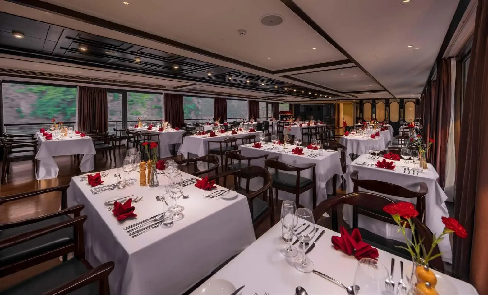 Interior of a ship dining room with rows of tables set with white tablecloths, red napkins, glasses and red carnations, with large windows showing green karst-like hillside outside.
