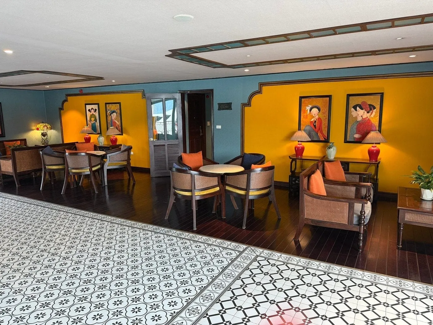 An indoor lounge with yellow and teal walls, rattan armchairs with orange cushions, round tables, red lamps, framed paintings of women in traditional dress, and patterned black-and-white tile flooring.