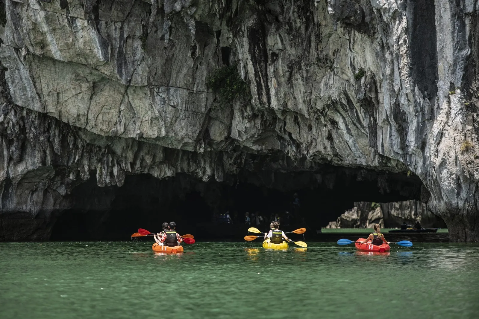 Three people kayaking in colorful kayaks through a limestone cave with green water.