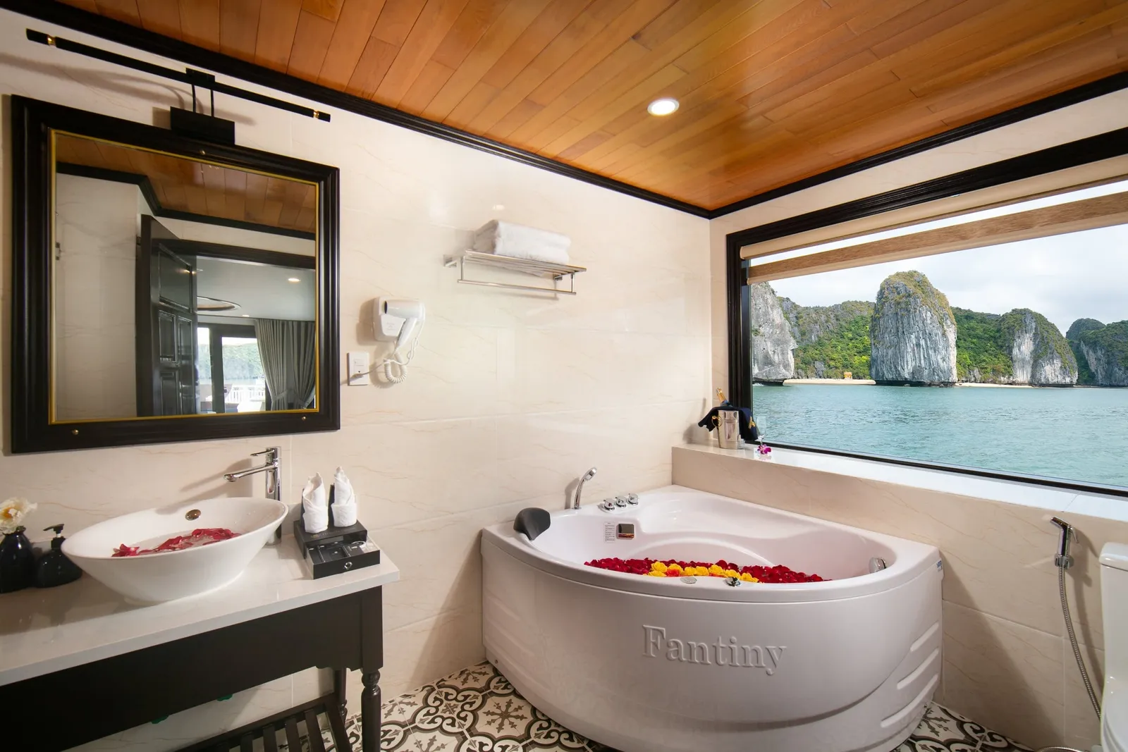 A bathroom with a corner jacuzzi tub filled with flower petals, a vessel sink, and a mirror, with a large window showing limestone karst islands and green water.