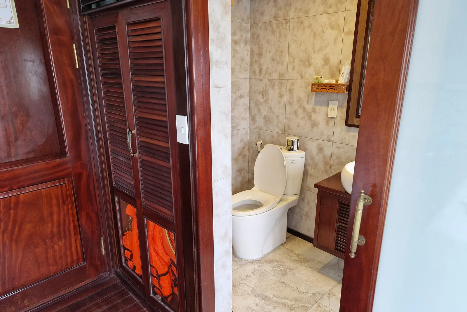 A boat cabin interior showing a bathroom with toilet and tiled walls, adjacent to dark wood cabinetry with orange life jackets stored inside.