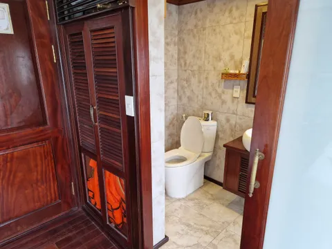 A boat cabin interior showing a bathroom with toilet and tiled walls, adjacent to dark wood cabinetry with orange life jackets stored inside.