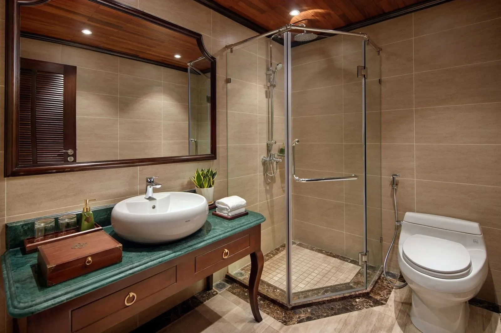 A bathroom with beige tiled walls, a glass corner shower, a white toilet, and a dark wood vanity with a green marble top, a white vessel sink, and a large mirror under a wooden ceiling.