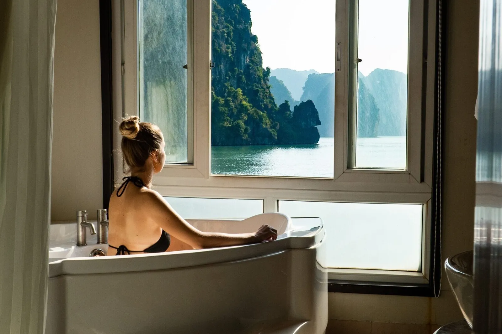 A person in a bathtub inside a cabin with windows showing limestone karst islands and calm water outside.