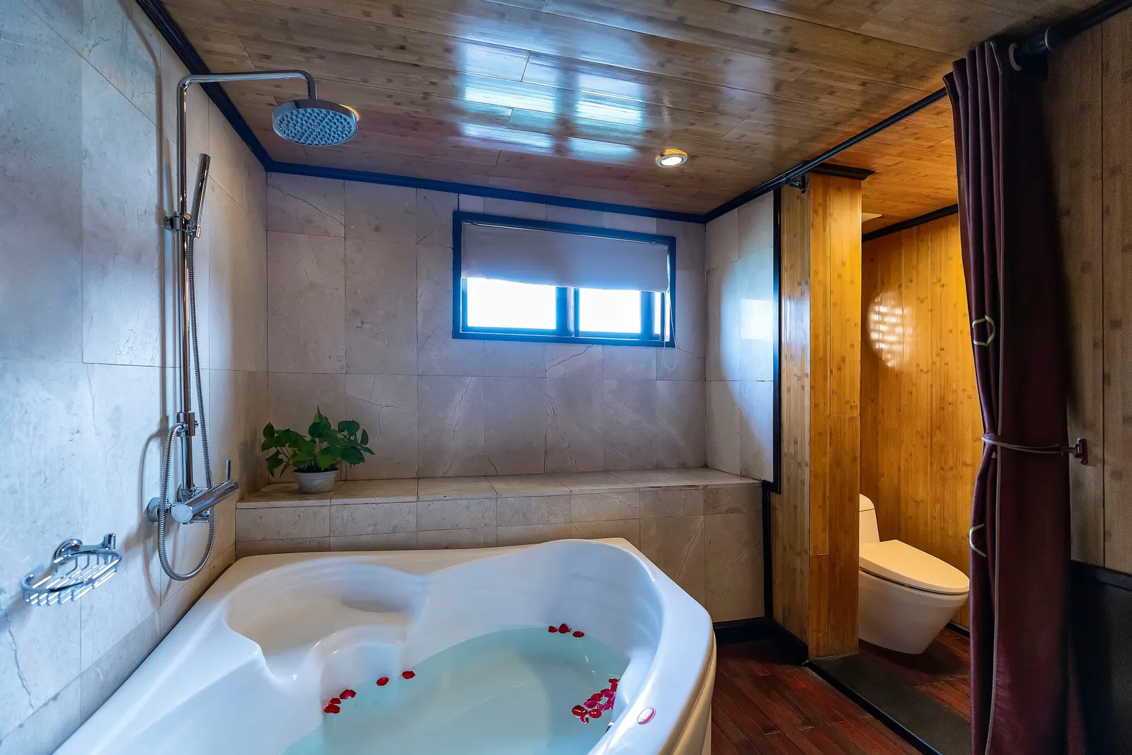 A bathroom with a corner bathtub filled with water and red petals, a rain shower head on a marble-tiled wall, a potted plant on a ledge, a window with a roller blind, a toilet behind a maroon curtain, and bamboo-style wood paneling on the ceiling and walls.