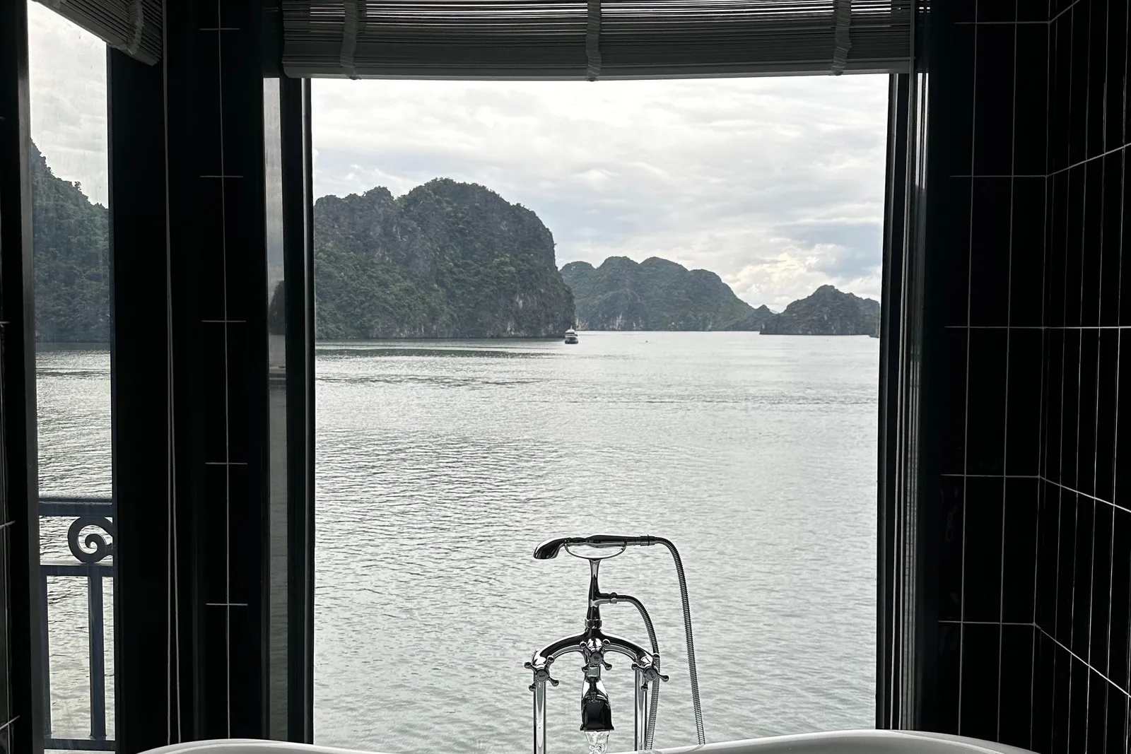A freestanding white bathtub filling with water sits in a dark-tiled bathroom, with a large window opening onto calm water and green limestone karst islands, and a small balcony railing visible at left.