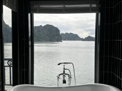 A freestanding white bathtub filling with water sits in a dark-tiled bathroom, with a large window opening onto calm water and green limestone karst islands, and a small balcony railing visible at left.
