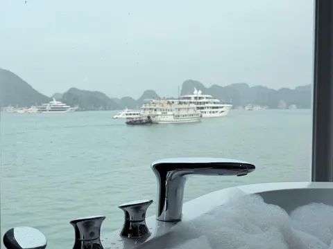 A bathtub with chrome fixtures in the foreground frames a view through a window of cruise ships and karst limestone islands in calm waters.