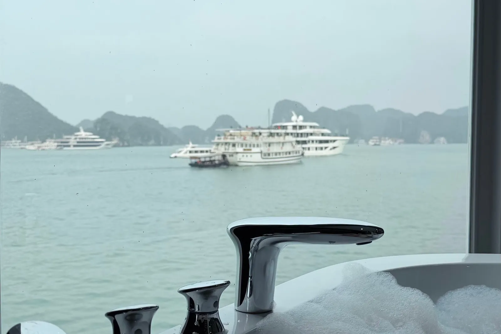 A bathtub with chrome fixtures in the foreground frames a view through a window of cruise ships and karst limestone islands in calm waters.