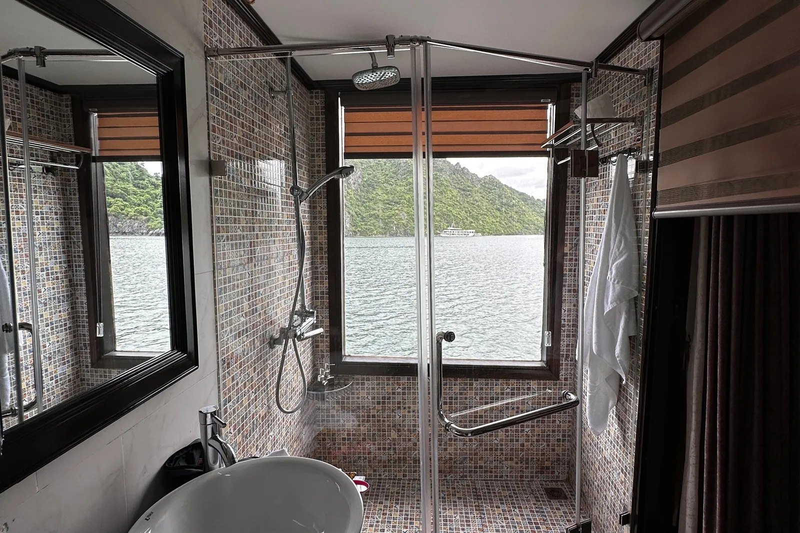 A modern boat bathroom with mosaic tile shower, vessel sink, and window view of green karst islands and water.