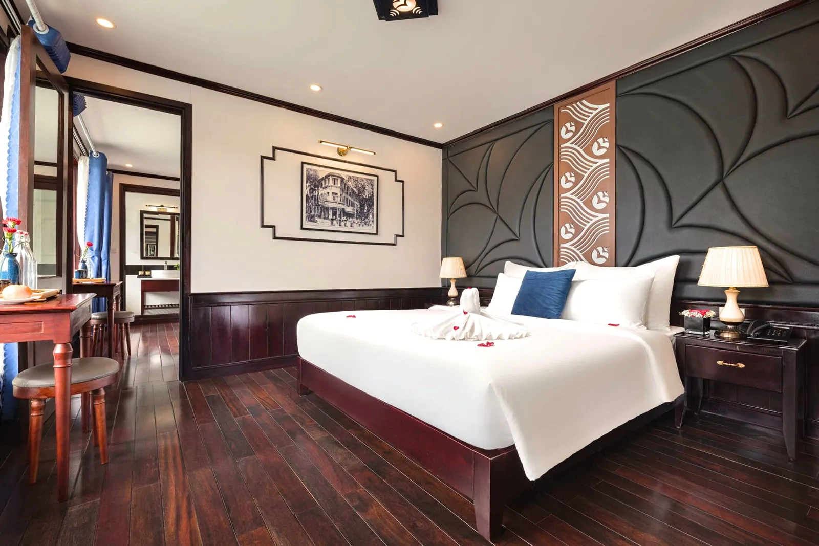 A bedroom interior with dark wood flooring, a bed with white linens and towel decoration, decorative wall panels, and a doorway to an ensuite bathroom.