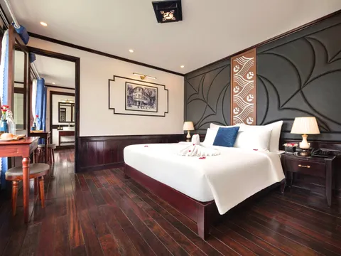 A bedroom interior with dark wood flooring, a bed with white linens and towel decoration, decorative wall panels, and a doorway to an ensuite bathroom.