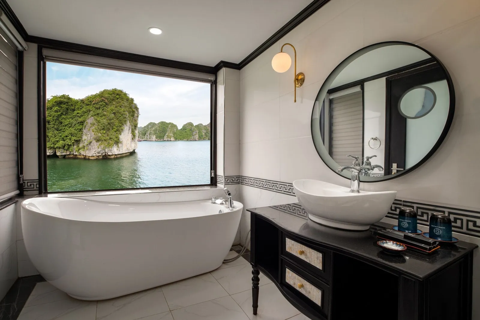 A bathroom with a freestanding white tub and a vanity with a vessel sink and round mirror, with a large window looking out on green karst islands and calm water.