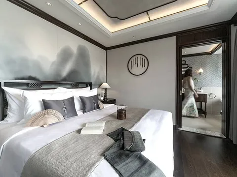 A dark-wood cabin with a large white bed, grey cushions, a folding fan and open book, an ink-mountain mural behind the headboard, and a blurred woman in a floral dress walking through the doorway to an en-suite bathroom.