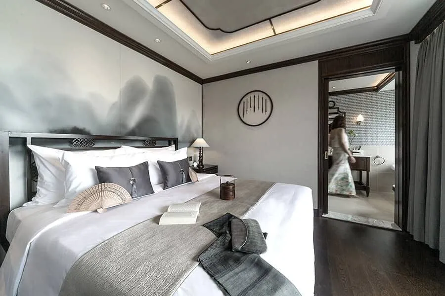 A dark-wood cabin with a large white bed, grey cushions, a folding fan and open book, an ink-mountain mural behind the headboard, and a blurred woman in a floral dress walking through the doorway to an en-suite bathroom.