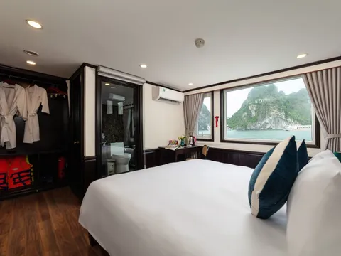 A cruise cabin with a white double bed, teal cushions, bathrobes and orange life jackets in an open wardrobe, a glass-walled bathroom, and a large window showing limestone karst islands on calm green water.