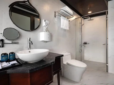 A marble-tiled bathroom with a vessel sink on a dark stone vanity, a round mirror, a wall-mounted hair dryer, a towel rack, a toilet, and a glass-enclosed rain shower.