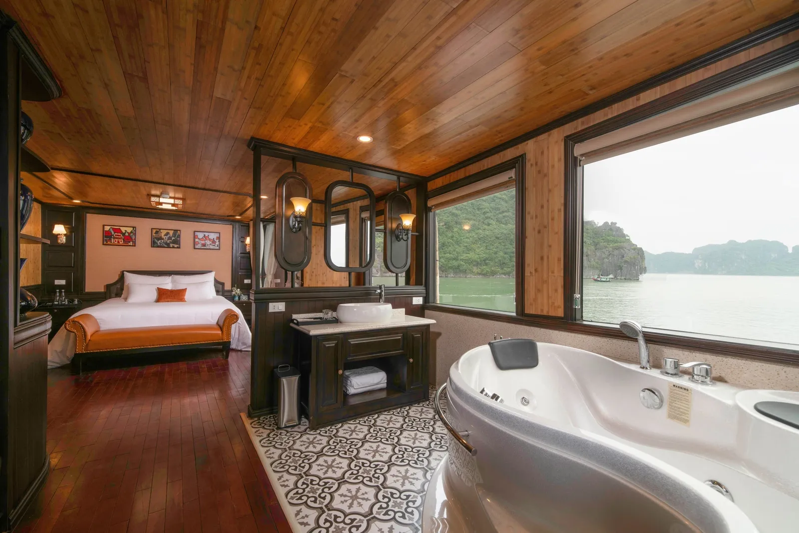 A luxury boat cabin interior with a bed, bathtub, and large windows showing limestone karsts and water outside.