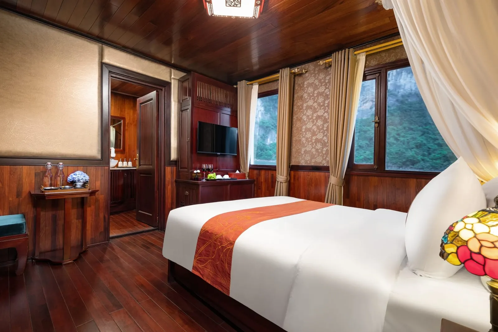 A dark-wood cruise cabin with a white double bed with orange runner, stained-glass lamp, TV, curtains, and windows showing forested limestone karst cliffs, with an open door to an ensuite bathroom.