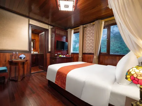 A dark-wood cruise cabin with a white double bed with orange runner, stained-glass lamp, TV, curtains, and windows showing forested limestone karst cliffs, with an open door to an ensuite bathroom.