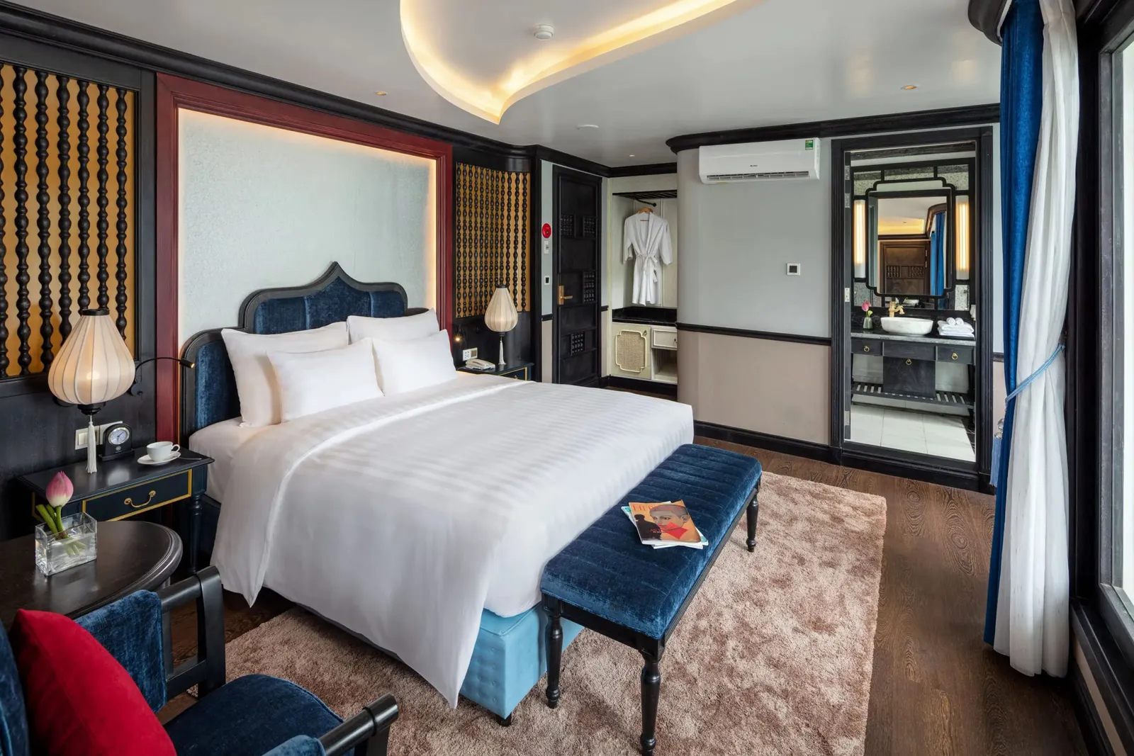 A cabin with a white-linen double bed, a blue velvet bench and headboard, silk lantern lamps, a pink lotus in a vase, and an open doorway to an en-suite bathroom, with a large window and curtains on the right.