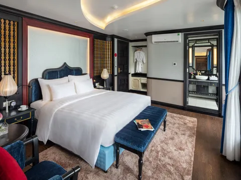 A cabin with a white-linen double bed, a blue velvet bench and headboard, silk lantern lamps, a pink lotus in a vase, and an open doorway to an en-suite bathroom, with a large window and curtains on the right.