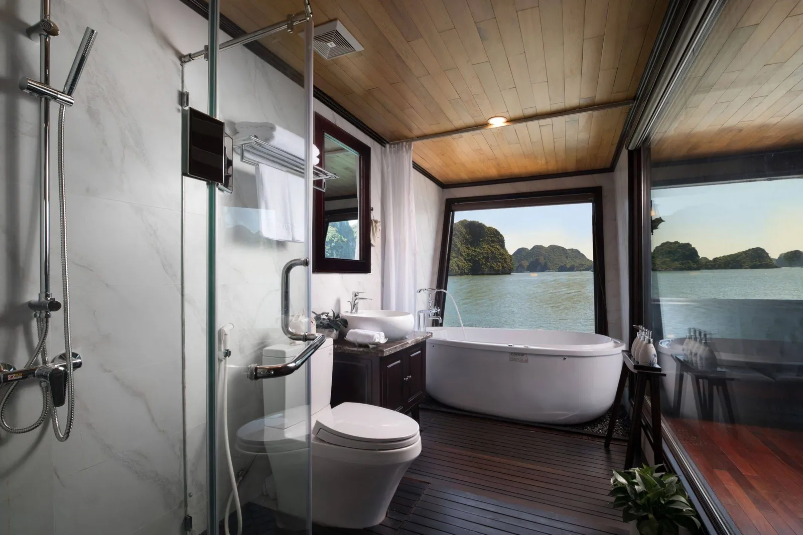 A cabin bathroom with a glass-enclosed shower, toilet, vanity and freestanding tub beneath a wooden ceiling, with a large window looking out onto calm water and green limestone karst islands.