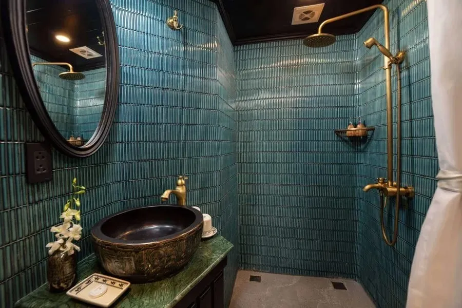 A bathroom with teal vertical tiles, a walk-in shower with brass fixtures, a dark ornate vessel sink on a green marble vanity, an oval black-framed mirror and a white orchid in a vase.