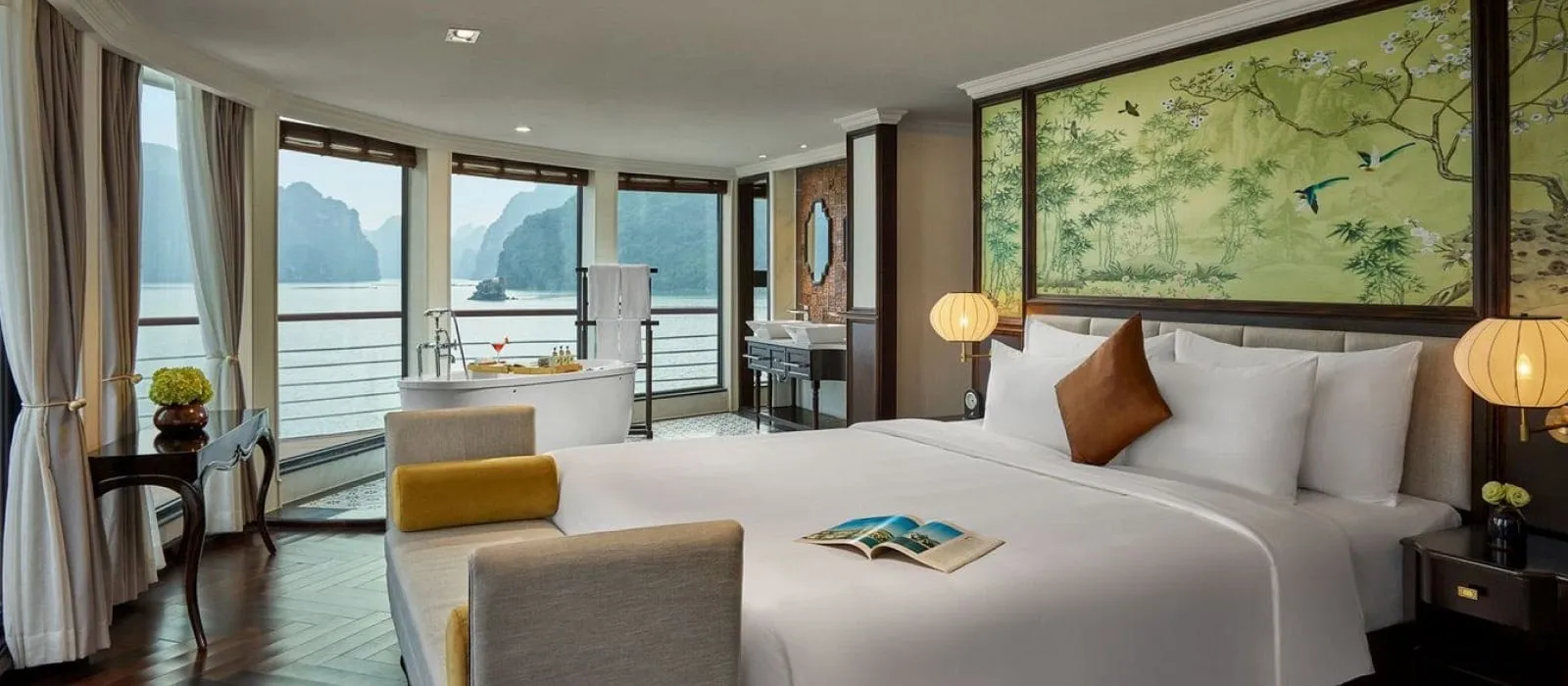 A cruise cabin with a large white bed, a freestanding bathtub by curved floor-to-ceiling windows, a green bird-and-branch painted panel, and limestone karsts over calm water outside.