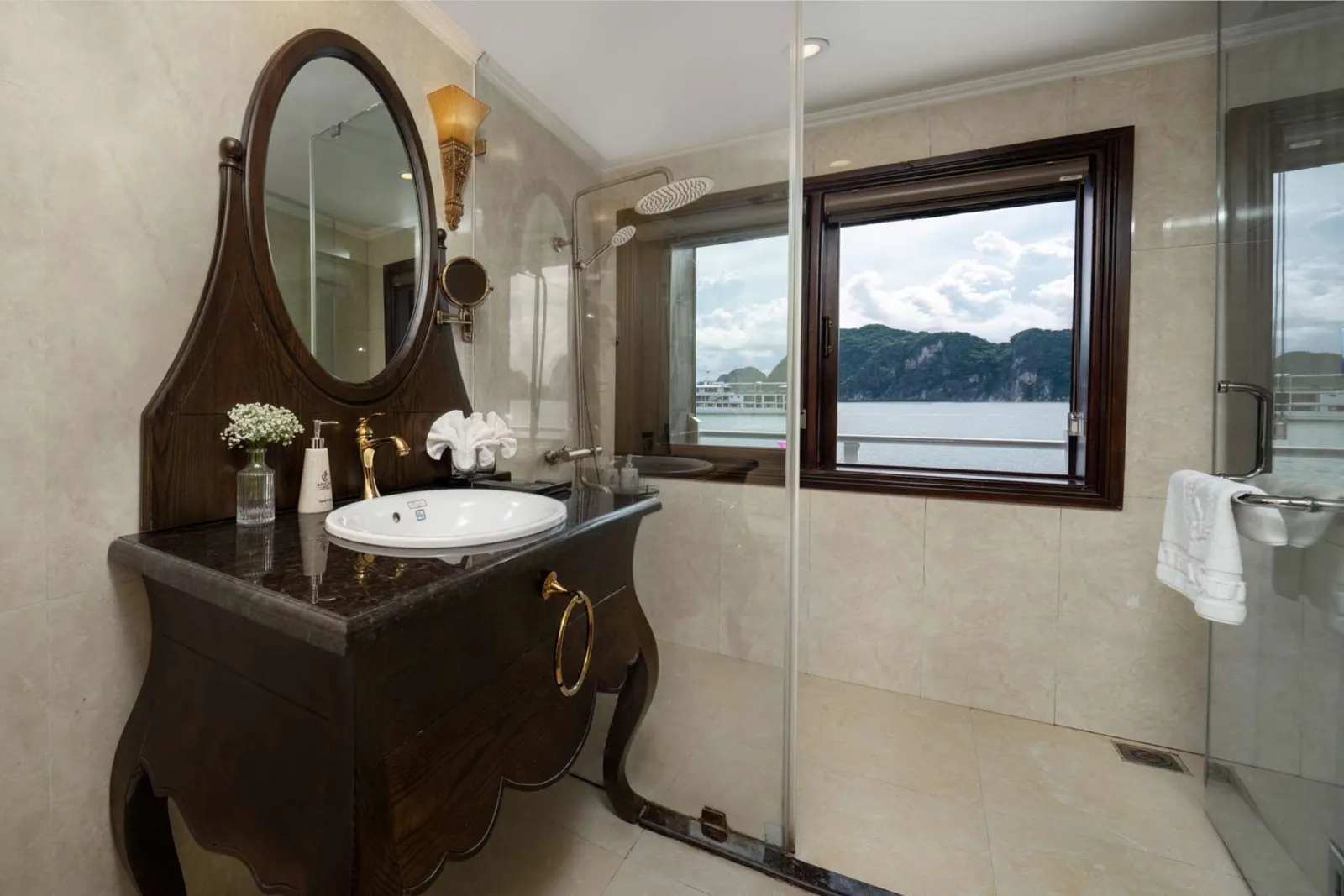 A bathroom with a dark wood vanity, oval mirror, round sink and glass shower, with a window showing limestone karst hills and calm water.
