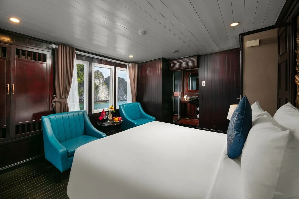 A dark-wood cruise cabin with a white double bed, blue pillow, two teal armchairs and a small table with fruit, with windows showing limestone karst cliffs over green water and an en-suite bathroom behind.