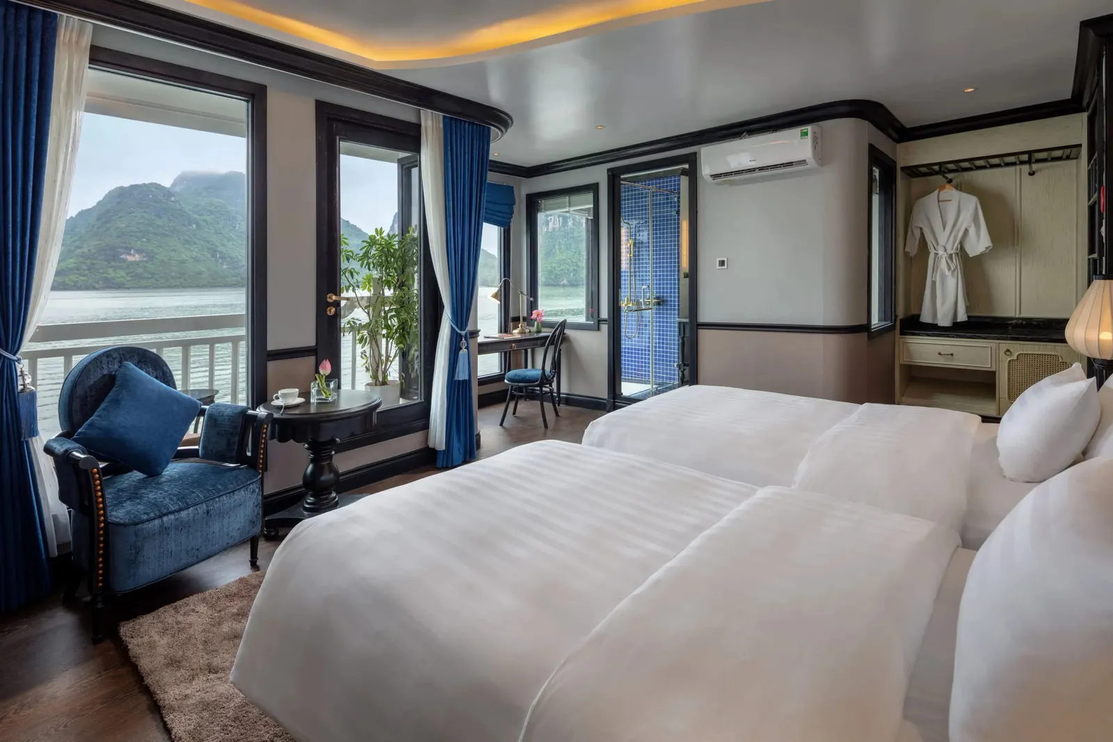 A cruise cabin with two white twin beds, a blue velvet armchair, a desk, a blue-tiled shower and a bathrobe, with windows and a balcony overlooking calm water and karst limestone hills.