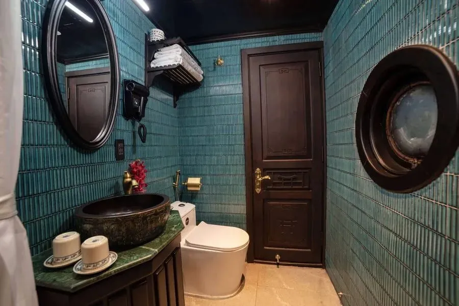 A bathroom with teal vertical tile walls, a dark wood door, a round porthole-style window, a dark bowl sink on a green marble counter with two cups on saucers, a white toilet, and towels on a wall shelf.